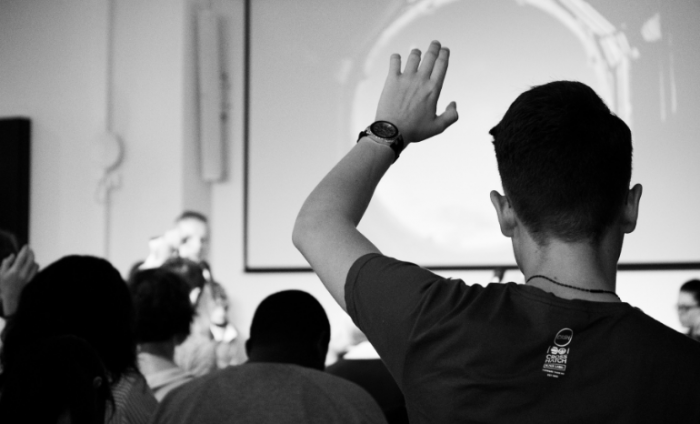 Black and white shot of an audience listening to a presentation and a man with his hand up to ask a question.